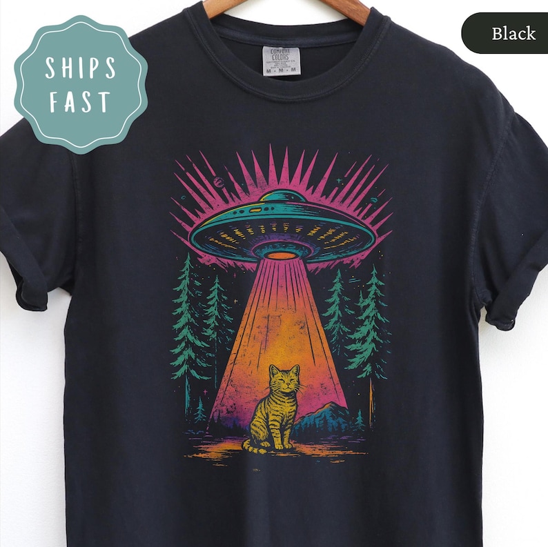 Op de afbeelding: Zwart T-shirt met een kleurrijke afbeelding van een UFO die een lichtstraal op een kat schijnt. Het ontwerp bevat een bos- en berglandschap met roze en turquoise tinten. Het shirt heeft een ronde hals.