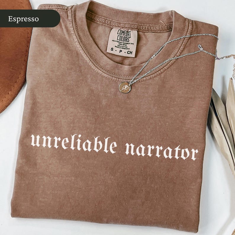 May include: Espresso-colored t-shirt with the words "unreliable narrator" in white gothic font. A silver necklace with a small pendant rests on the shirt. The shirt has a Comfort Colors tag.