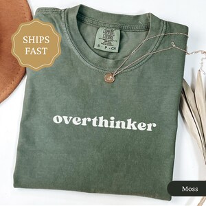 May include: A moss green t-shirt with the word "overthinker" printed in white. A gold necklace with a pendant rests on the shirt. The shirt is made by Comfort Colors.