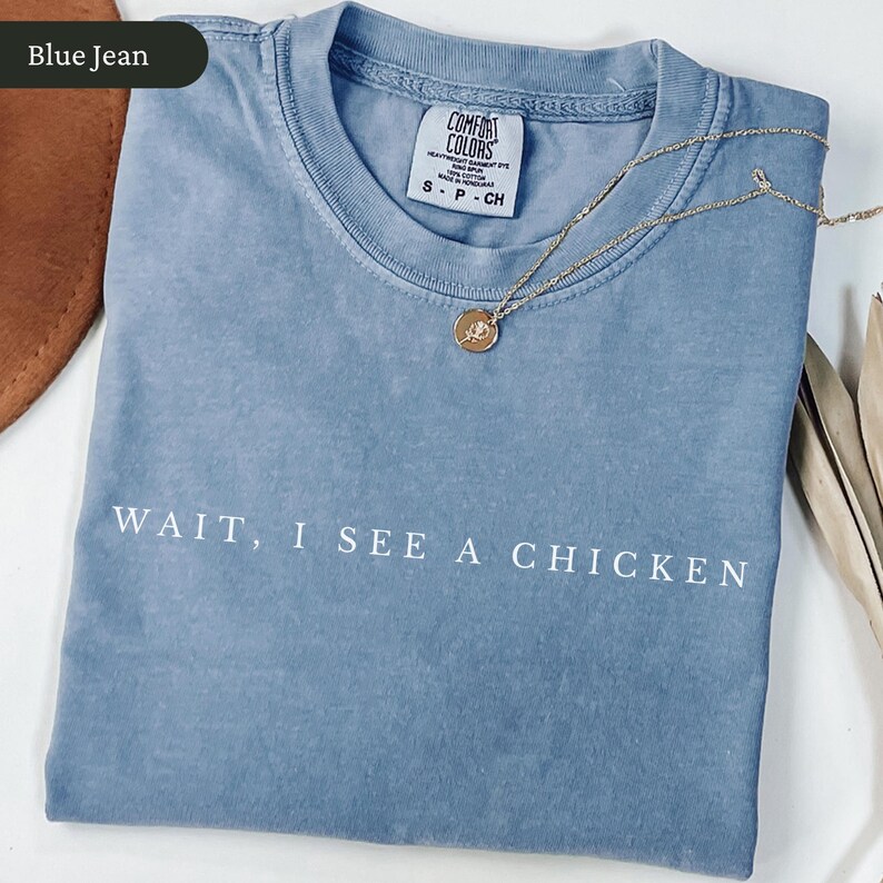 May include: A blue Comfort Colors t-shirt with the text "WAIT, I SEE A CHICKEN" in white. A gold necklace with a pendant rests on the shirt. The shirt is folded and has a tag at the neckline.