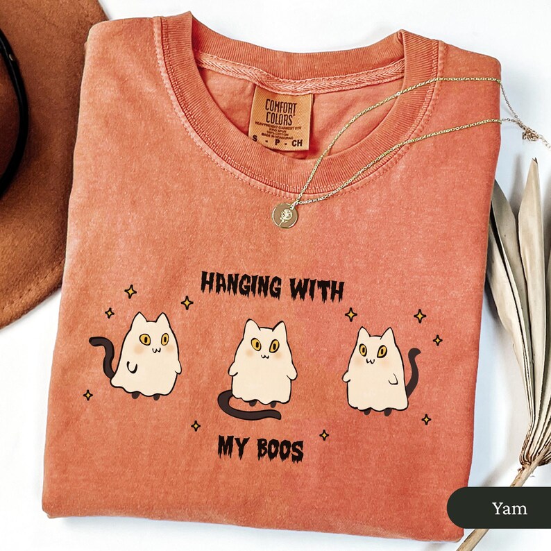 May include: A coral-coloured Comfort Colors t-shirt with the phrase "Hanging with my Boos" and three cartoon ghost cats. A gold necklace with a pendant is draped on the shirt. The shirt is folded on a white surface.