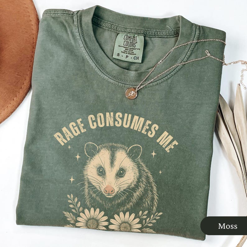 May include: A moss green t-shirt featuring the text "RAGE CONSUMES ME" and an opossum illustration with flowers and stars. The shirt has a Comfort Colors tag. A gold necklace with a pendant is draped on the shirt.