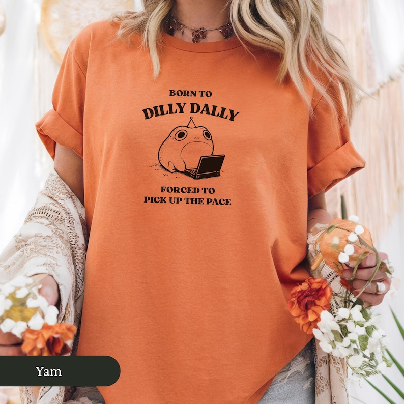 May include: Orange t-shirt with rolled-up sleeves featuring the text "BORN TO DILLY DALLY FORCED TO PICK UP THE PACE" and a cartoon frog with a laptop. The shirt is a solid orange color.
