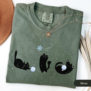 Puede incluir: Camiseta verde musgo con un dise&ntilde;o juguet&oacute;n de tres gatos negros interactuando con un copo de nieve y bolas de nieve. La camiseta tiene cuello redondo y mangas cortas. Un delicado collar con un colgante est&aacute; sobre la camiseta.