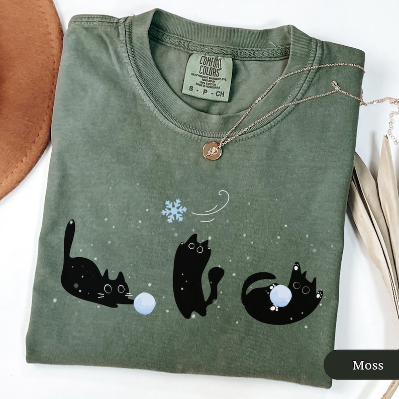 Puede incluir: Camiseta verde musgo con un dise&ntilde;o juguet&oacute;n de tres gatos negros interactuando con un copo de nieve y bolas de nieve. La camiseta tiene cuello redondo y mangas cortas. Un delicado collar con un colgante est&aacute; sobre la camiseta.