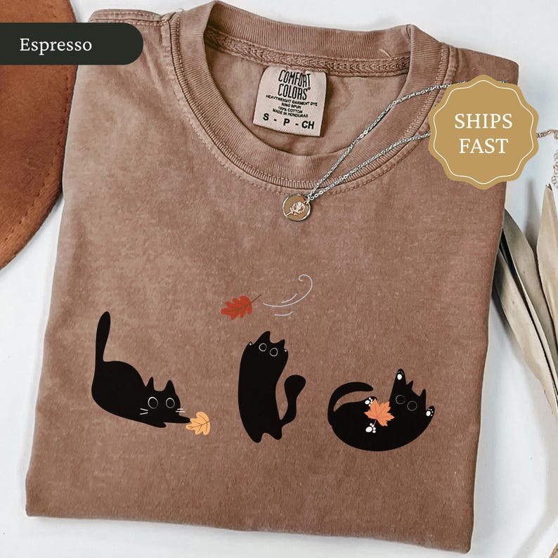 May include: Espresso-colored t-shirt with a playful design of three black cats interacting with autumn leaves. The shirt features a Comfort Colors label and a silver necklace with a pendant. The text "SHIPS FAST" is also visible.