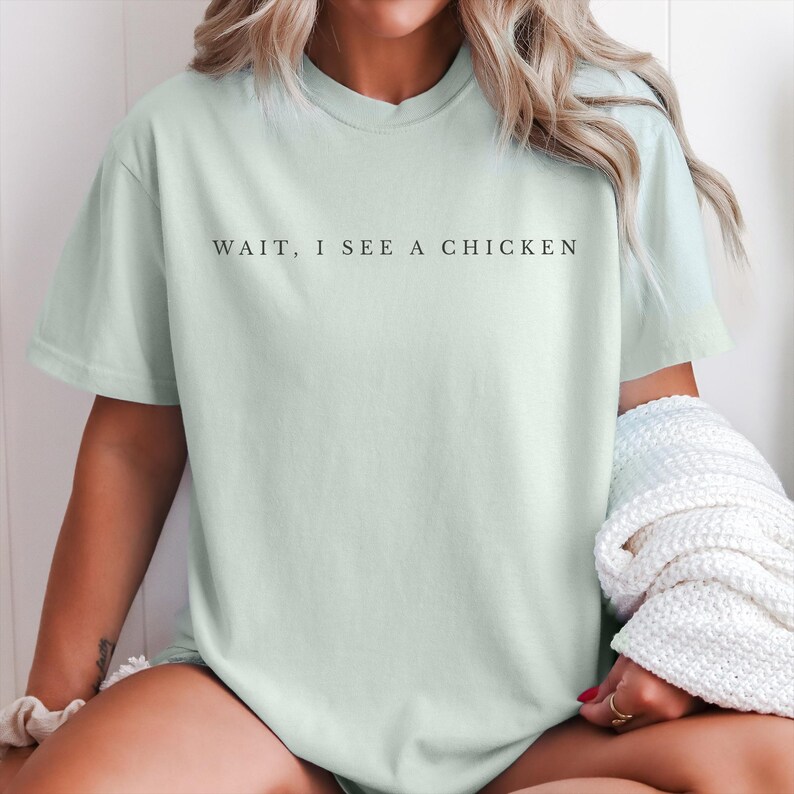 May include: A pale green t-shirt featuring the black text "WAIT, I SEE A CHICKEN". The top has a crew neck and short sleeves. A white knitted jumper is draped over the shoulders.