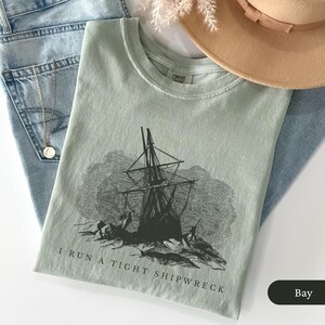 May include: A light green t-shirt with a black graphic of a ship in a storm, with the text "I RUN A TIGHT SHIPWRECK". The shirt is folded on top of blue jeans, with a necklace and a tan hat visible.