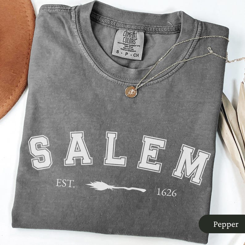 Peut inclure: Un t-shirt gris Comfort Colors avec le mot "SALEM" en lettres blanches. En dessous du mot "SALEM" se trouve le texte "EST. 1626" avec un motif de balai. Un collier en or avec un pendentif est drap&eacute; sur le t-shirt.