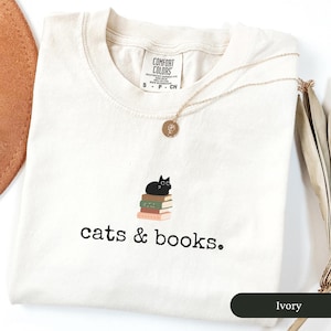 Może przedstawiać: Koszulka w kolorze kości słoniowej z czarnym kotem siedzącym na stosie książek. Pod kotem nadrukowany jest tekst "cats & books."