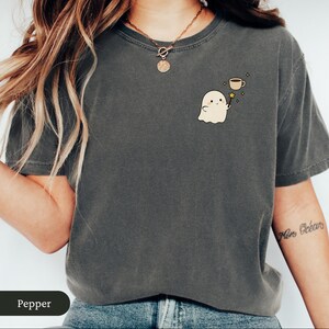 May include: Dark grey t-shirt featuring a cartoon ghost holding a magic wand with a coffee cup at the end. The ghost is beige with a smiling face. The shirt is a crew neck and has short sleeves.
