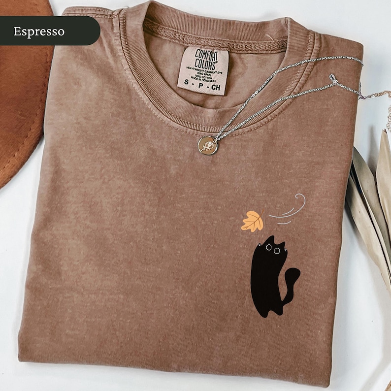 May include: Espresso-coloured t-shirt featuring a black cat graphic and a falling leaf design. The shirt has a crew neck and short sleeves. A silver necklace with a small pendant is draped across the shirt. The shirt is labelled "Comfort Colors".