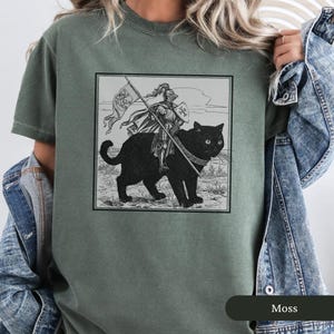 May include: A green t-shirt with a black and white illustration of a knight riding a black cat. The knight is wearing armor and holding a sword. The cat is wearing a collar. The illustration is in a square frame.