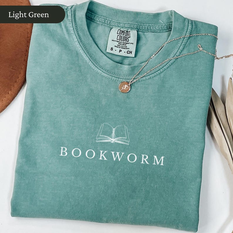 Op de afbeelding: Lichtgroen T-shirt met het woord "BOOKWORM" in het wit gedrukt, samen met een illustratie van een open boek. Een zilveren ketting met een hanger ligt op het shirt. Het shirt is opgevouwen en heeft een label met "Comfort Colors".