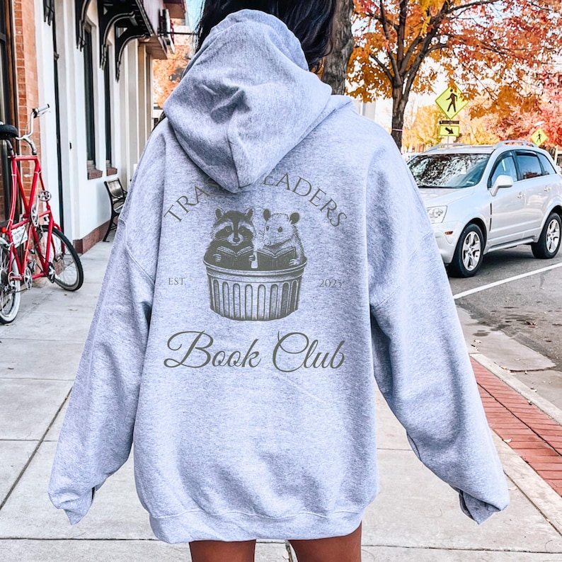 May include: A light gray hoodie with the text "Trail Readers Book Club" in a stylized font. The back of the hoodie features a graphic of a raccoon and a hamster reading a book. The hoodie has a drawstring hood.