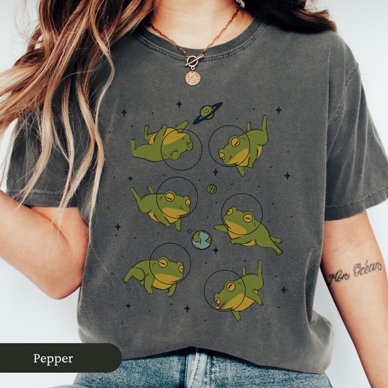 May include: Dark grey t-shirt featuring a whimsical design of green frogs in space suits floating among planets and stars. The shirt has a crew neck and short sleeves. The word "Pepper" is printed at the bottom of the shirt.