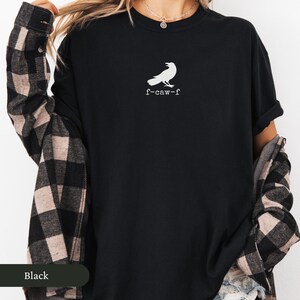 May include: Black t-shirt with a white raven graphic and the text "f-caw-f". The shirt has short sleeves and a crew neck. A plaid shirt is draped over the shoulders.