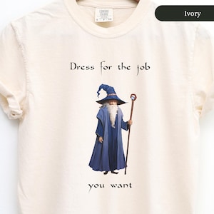 May include: Ivory t-shirt featuring a graphic of a wizard wearing a blue robe and holding a staff. The text "Dress for the job you want" is printed on the shirt.