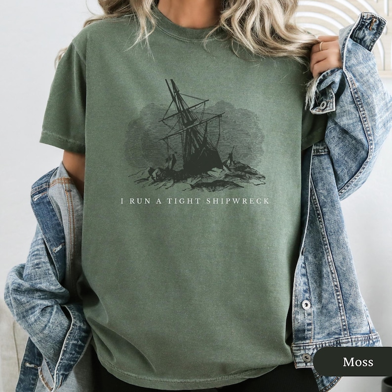May include: Moss green t-shirt featuring a black and white illustration of a shipwreck. The text "I RUN A TIGHT SHIPWRECK" is printed below the graphic. The shirt has short sleeves and a crew neck.