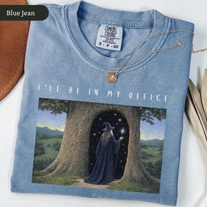 Op de afbeelding: Blauw spijkerbroekkleurig T-shirt met de tekst "I'll Be In My Office". De afbeelding toont een tovenaar in een blauwe jurk en hoed die in een boomdeur staat. Een gouden ketting met een hanger ligt op het shirt.