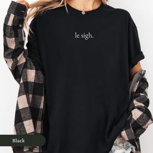 Pu&ograve; includere: T-shirt nero girocollo con la scritta "le sigh." in bianco. La modella indossa una camicia di flanella a quadri neri e beige sopra la t-shirt. La parola "Black" &egrave; visibile in un riquadro verde scuro in basso a sinistra.