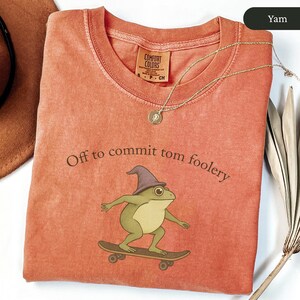 Puede incluir: Una sudadera Comfort Colors color &ntilde;ame con la frase "Off to commit tom foolery" impresa en la parte delantera. El gr&aacute;fico presenta una rana de dibujos animados con un sombrero de mago y patinando. Un collar de oro con un colgante est&aacute; sobre la camisa.