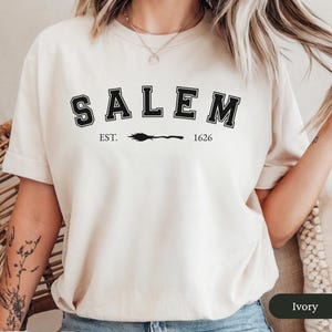 Peut inclure: T-shirt de couleur ivoire avec le mot "SALEM" en lettres noires, soulign&eacute; de blanc. En dessous, le texte "EST. 1626" avec un dessin de balai. Le t-shirt est &agrave; col rond.