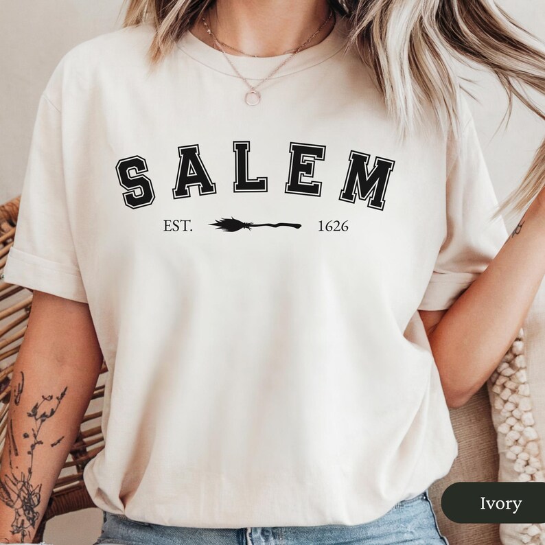 Peut inclure: T-shirt de couleur ivoire avec le mot "SALEM" en lettres noires, soulign&eacute; de blanc. En dessous, le texte "EST. 1626" avec un dessin de balai. Le t-shirt est &agrave; col rond.