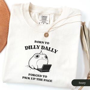 May include: Ivory-colored t-shirt with the text "BORN TO DILLY DALLY FORCED TO PICK UP THE PACE" and a cartoon character with a laptop. A gold necklace with a pendant is draped across the shirt.