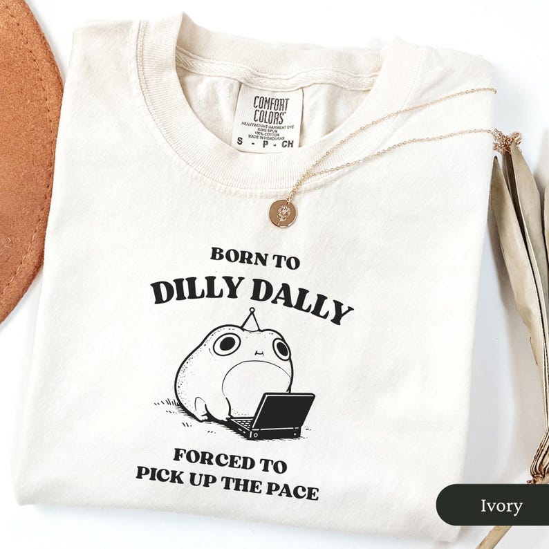 May include: Ivory-colored t-shirt with the text "BORN TO DILLY DALLY FORCED TO PICK UP THE PACE" and a cartoon character with a laptop. A gold necklace with a pendant is draped across the shirt.