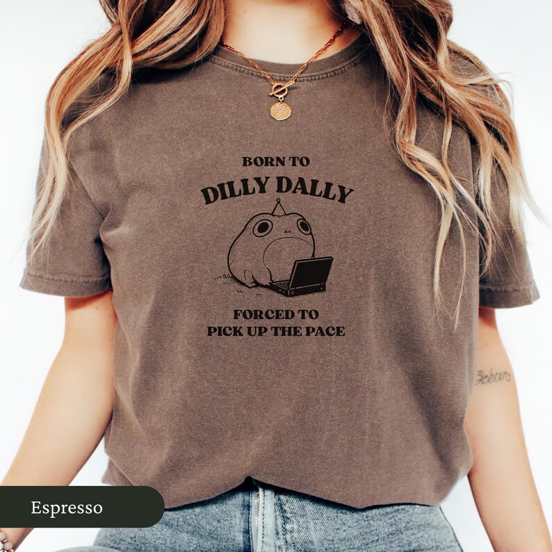 May include: Espresso-colored t-shirt featuring the text "BORN TO DILLY DALLY, FORCED TO PICK UP THE PACE" above a cartoon frog with a laptop. The shirt has short sleeves and a crew neck.