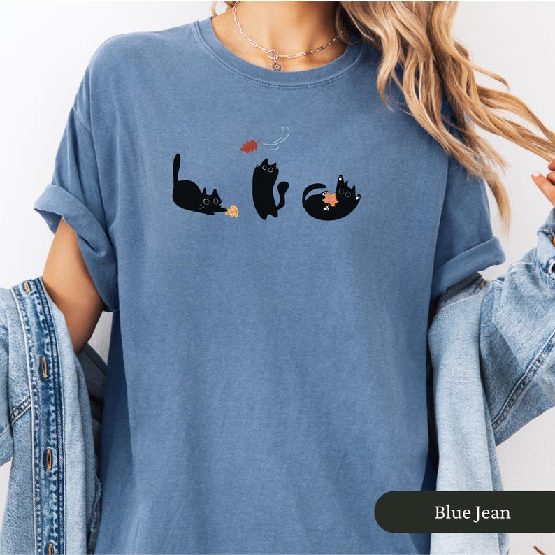 May include: Blue jean colored t-shirt featuring a playful graphic of three black cats interacting with autumn leaves. The cats are in different poses, with one reaching for a leaf. The shirt has a relaxed fit and rolled sleeves.