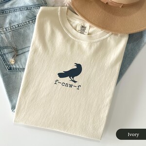 May include: Ivory-colored t-shirt with a navy blue crow graphic and the text "f-caw-f". The shirt is folded and placed on top of blue jeans, with a necklace and a tan hat in the background. The shirt is labeled "Comfort Colors".