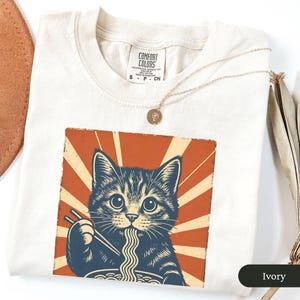 May include: Ivory-colored t-shirt featuring a graphic of a cat eating ramen with chopsticks, set against a retro orange and beige sunburst design. The shirt has a crew neck and short sleeves. A gold necklace with a pendant is draped on the shirt.