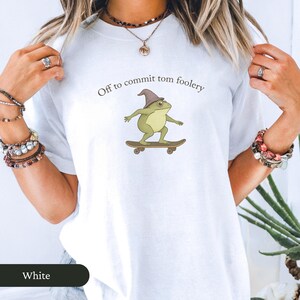 Puede incluir: Camiseta blanca con una rana de dibujos animados en monopat&iacute;n, con un sombrero de mago. El texto "Off to commit tom foolery" est&aacute; impreso encima de la rana. Un estilo casual.