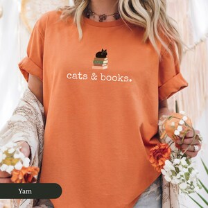 Może przedstawiać: Pomarańczowy T-shirt z czarnym kotem siedzącym na stosie książek. Na koszulce nadrukowany jest napis "cats & books."
