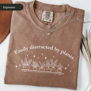 May include: Espresso-coloured t-shirt featuring the text "Easily distracted by plants" and a graphic of potted plants. A silver necklace with a small pendant rests on the shirt. The shirt is folded on a white surface.