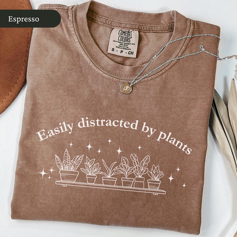 May include: Espresso-coloured t-shirt featuring the text "Easily distracted by plants" and a graphic of potted plants. A silver necklace with a small pendant rests on the shirt. The shirt is folded on a white surface.