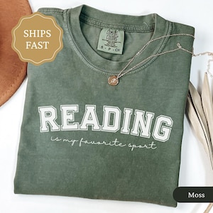 May include: Moss green t-shirt with the text "READING is my favorite sport" in white block letters. A gold necklace with a pendant rests on the shirt. The shirt is folded and displayed on a white surface.