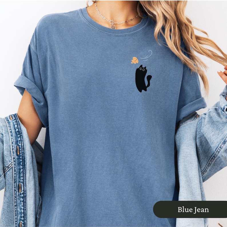 May include: Blue jean coloured t-shirt with rolled sleeves. A black cat graphic is on the left chest, reaching for a falling leaf. The shirt is a relaxed fit, with the words "Blue Jean" in a dark green rectangle at the bottom.