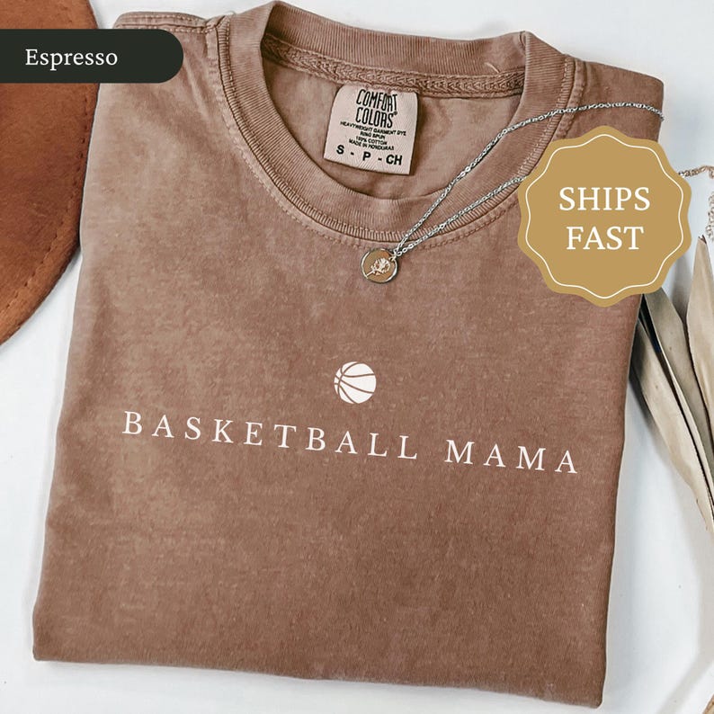 以下が含まれることがあります： 「BASKETBALL MAMA」の文字とバスケットボールのグラフィックが入ったエスプレッソカラーのTシャツ。クルーネックと半袖です。ペンダント付きのシルバーネックレスがTシャツの上にかけられています。