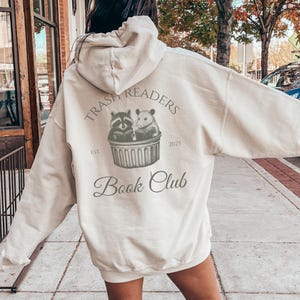 May include: A cream-colored hoodie with a graphic that reads "Trash Readers Book Club." The design features a raccoon and a hamster reading books in a trash can. The text "Est. 2025" is also included.