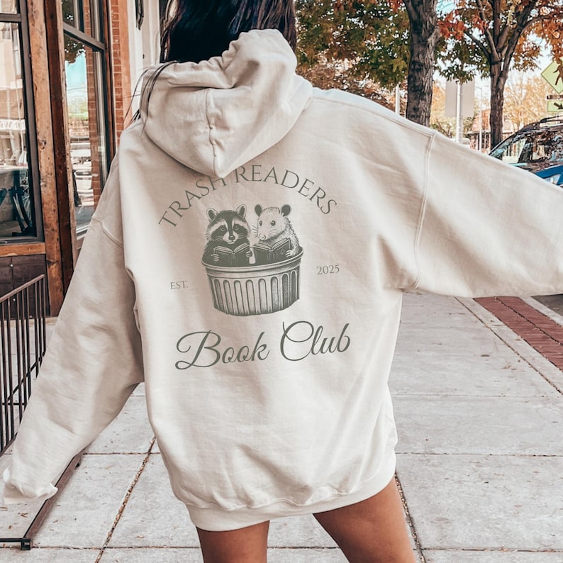 May include: A cream-colored hoodie with a graphic that reads "Trash Readers Book Club." The design features a raccoon and a hamster reading books in a trash can. The text "Est. 2025" is also included.