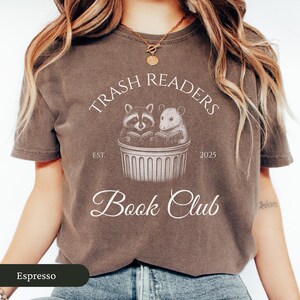 Puede incluir: Una camiseta marr&oacute;n con un dise&ntilde;o gr&aacute;fico blanco que representa un mapache y una rata leyendo libros en un contenedor de basura. El texto dice "Trash Readers Est. 2025 Book Club".