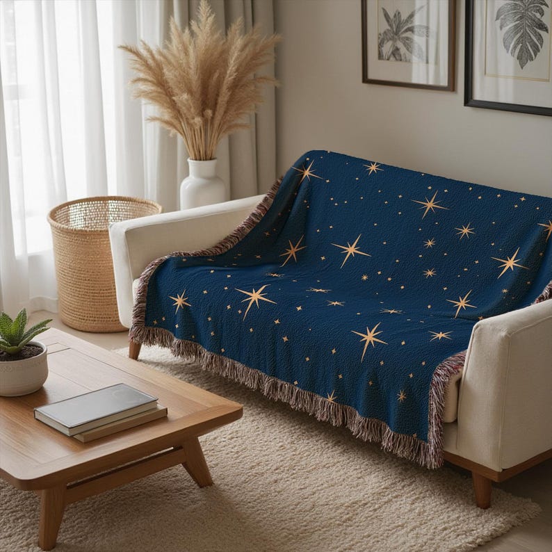 Puede incluir: Una manta azul marino con un estampado de estrellas doradas sobre un sof&aacute; blanco. La manta tiene un borde con flecos y est&aacute; colocada en un sal&oacute;n con una mesa de centro de madera y una cesta de mimbre.