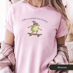 Puede incluir: Camiseta rosa claro con la frase "Off to commit tom foolery" y una rana de dibujos animados con sombrero de mago en monopat&iacute;n. La rana es verde y amarilla. La camiseta es de color liso.
