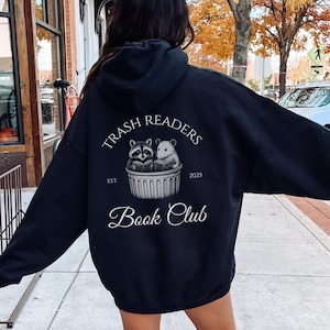 Peut inclure: Sweat à capuche noir avec l'inscription "Trash Readers Book Club". Le motif représente un raton laveur et une souris lisant des livres dans une poubelle. Le texte "Est. 2025" est également inclus. Style décontracté.