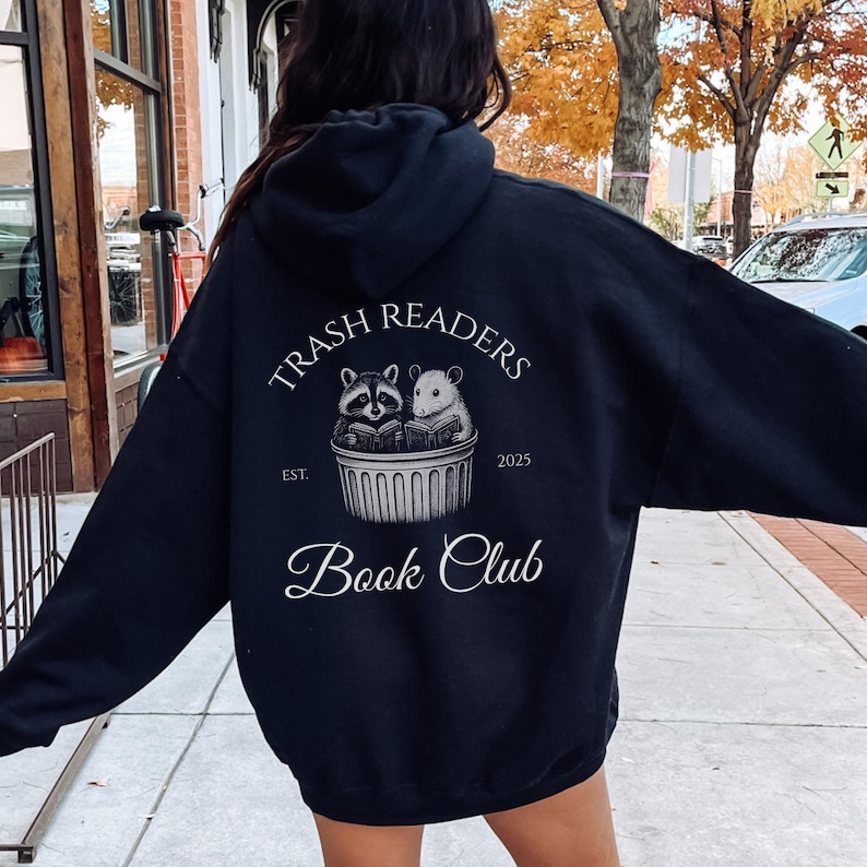 May include: Black hoodie with the text "Trash Readers Book Club." The design features a raccoon and a mouse reading books in a trash can. The text "Est. 2025" is also included. The hoodie is a casual style.