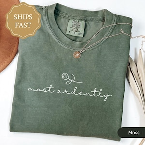 Peut inclure: Un t-shirt vert mousse plié avec l'inscription blanche "most ardently" et un motif floral. Un collier doré avec un pendentif repose sur le t-shirt. L'étiquette du t-shirt indique "Comfort Colors". La couleur est "Moss".