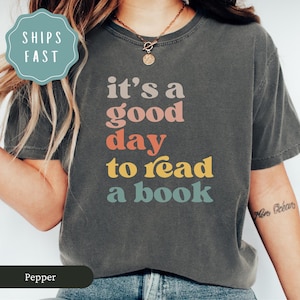 May include: Dark grey t-shirt featuring the phrase "it's a good day to read a book" in a retro font. The text is in shades of pink, orange, yellow, and teal. The shirt has a crew neck and a relaxed fit.